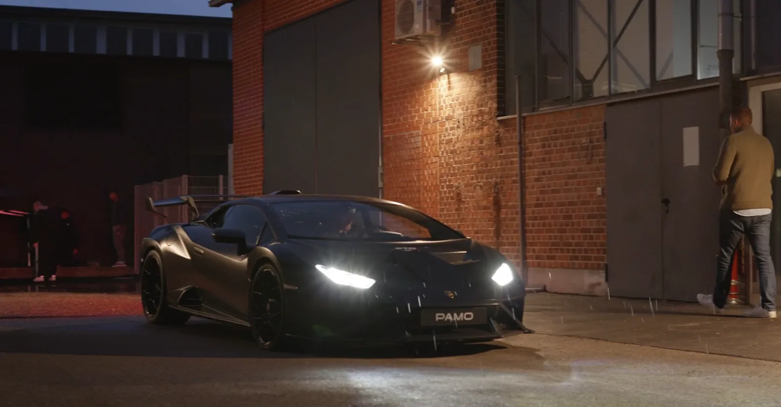 Schwarzer Supersportwagen bei Nacht vor einer Backsteinfassade, Crew am Rand im Regen