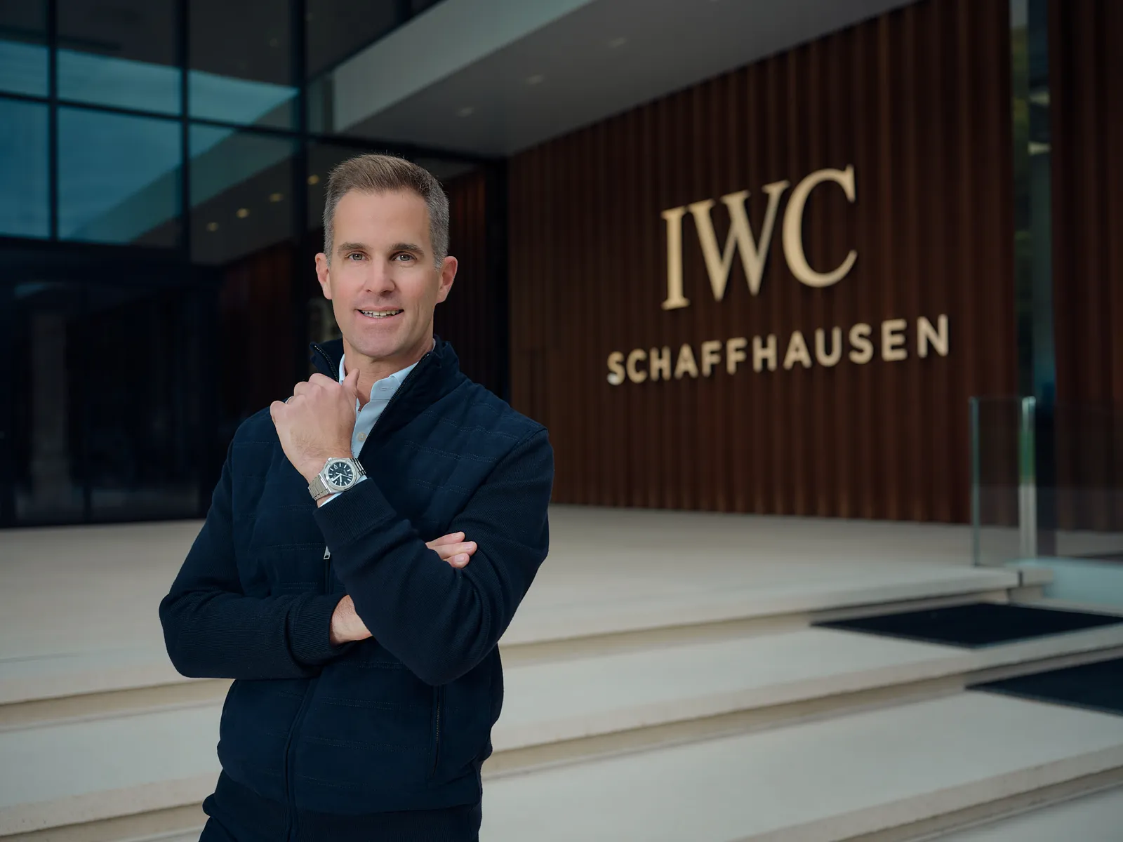 Mann in dunkelblauer Strickjacke zeigt seine Armbanduhr vor dem goldenen IWC Schriftzug am Eingang