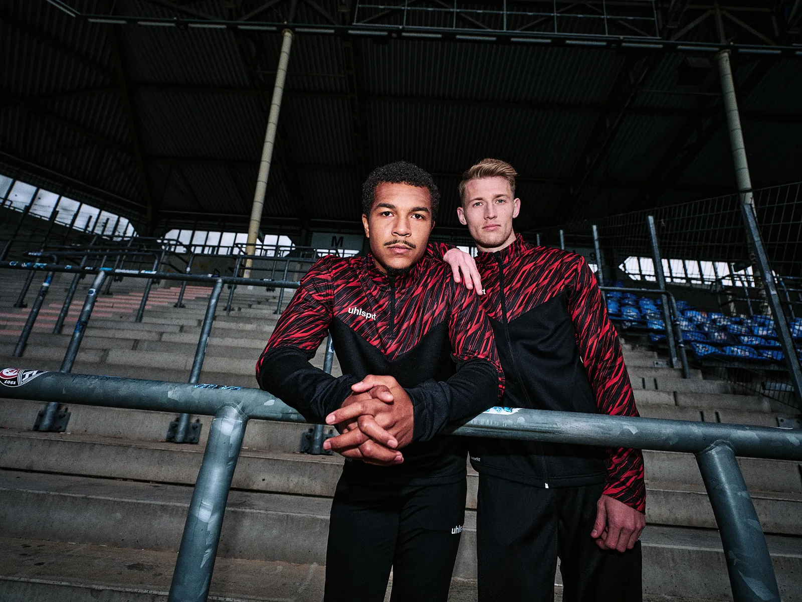 Zwei Spieler in schwarz-roten uhlsport-Trainingsjacken in der dunklen Tribünenhalle